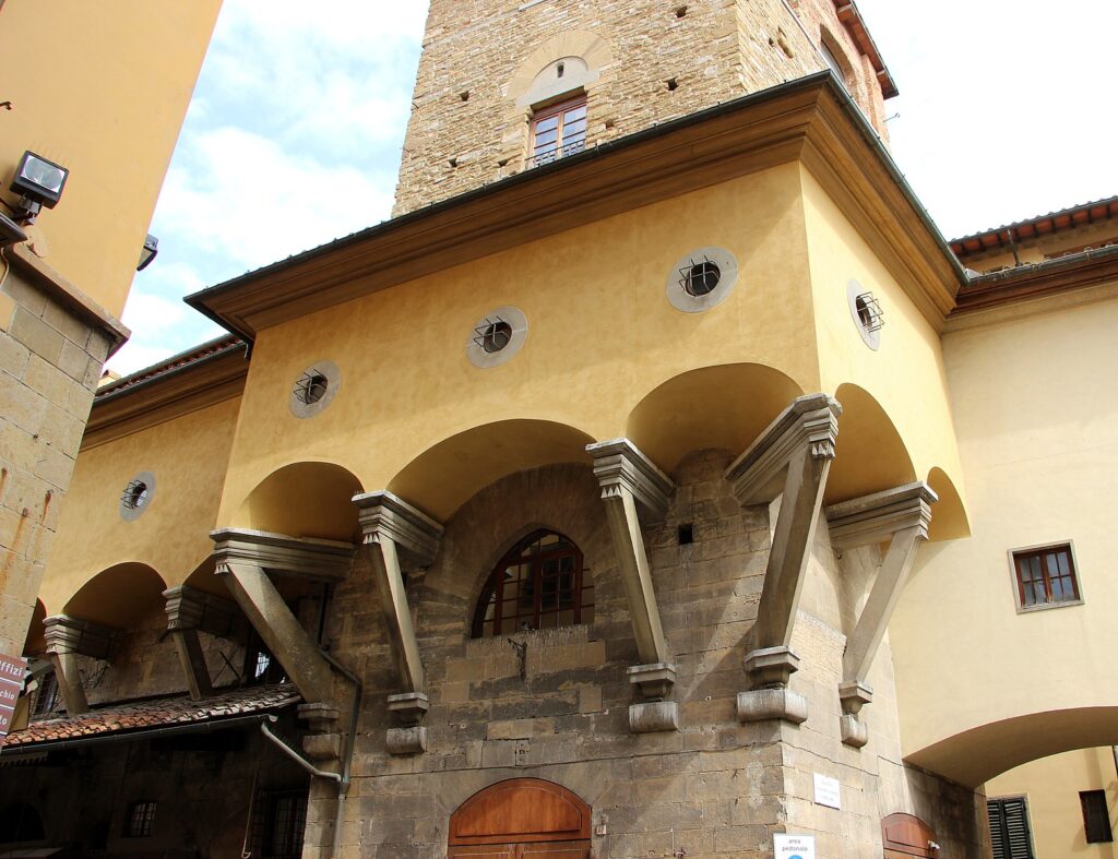Photograph of a building in Florence. A tall thin stone section rises up, from street level several stories. About the level of the second story, a yellow section sticks out from the outside of it, awkwardly wrapping around the outside of the stone part, supported by elegant sticky - outy triangular struts. The yellow section has several small circular windows, much too small for a human to climb through, barely large enough for a chubby cat.